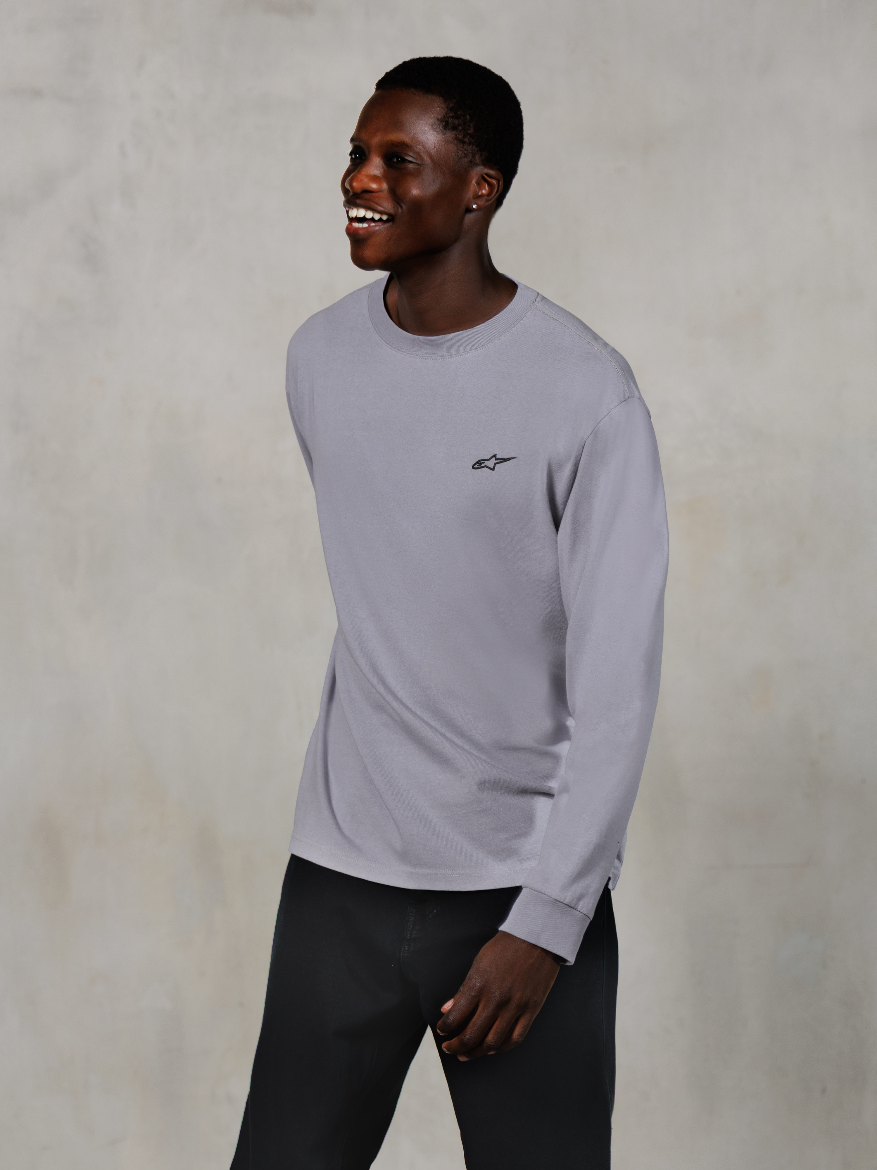 Alpinestars Essentials Ageless LS Tee, koszulka z długim rękawem, srebrna, małe czarne logo na piersi, noszona przez uśmiechniętego modela w ciemnych spodniach, swobodny nowoczesny krój, codzienna odzież lifestylowa w stylu motorsportowym