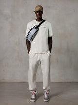 Alpinestars Essentials Ageless SS Tee, t-shirt z krótkim rękawem, kolor złamanej bieli, z małym czarnym logo na piersi, noszony z pasującymi joggersami, beżową czapką z daszkiem i szarą torbą typu crossbody, casualowy styl streetwear