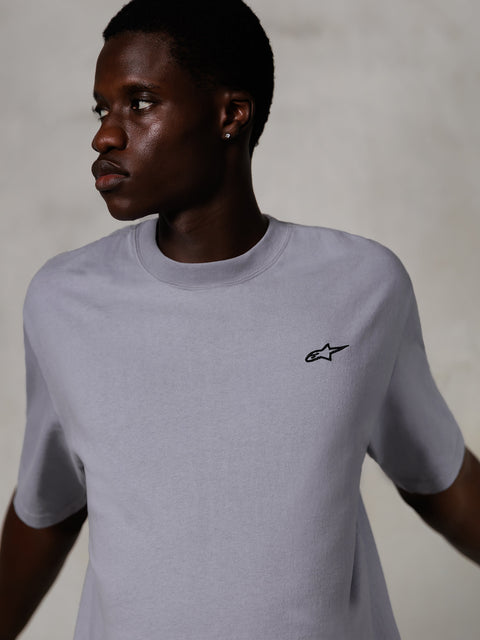 Model w koszulce Alpinestars Essentials Ageless SS Tee, srebrny T-shirt z krótkim rękawem, małe czarne logo na piersi, luźny krój, czysty i minimalistyczny design streetwearowy