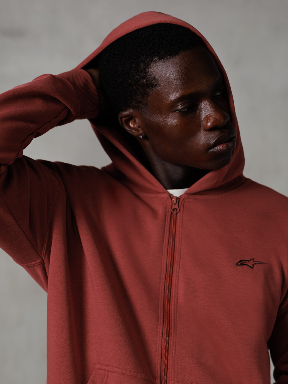 Alpinestars Essentials Ageless Zip Hoodie, ceglasta czerwień, codzienna odzież streetwearowa, założony kaptur i dłoń za głową, małe czarne logo Astars na piersi, polarowa bluza o luźnym kroju