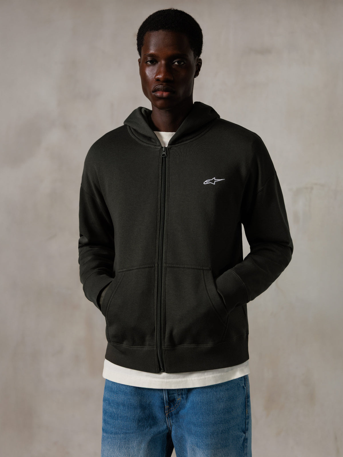 Alpinestars Essentials Ageless Zip Hoodie, bluza z kapturem w kolorze Off Black, rozpinana na zamek, małe białe logo Alpinestars na piersi, luźny krój, model w niebieskich dżinsach i białym t-shircie, streetwear inspirowany sportami motorowymi