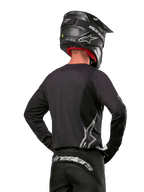 Motocross rider viewed from the back wearing the Alpinestars 2024 Fluid Graphite Jersey in Black Silver, featuring a large silver logo on the side, paired with a black carbon-texture helmet and black pants, professional off-road racing gear