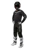 Alpinestars 2024 Fluid Graphite Gear Set, kompletny strój do wyścigów motocrossowych, czarny z szarymi i białymi logotypami, obejmujący koszulkę i spodnie noszone z dopasowanym czarnym kaskiem oraz biało-czarnymi rękawicami i butami, zaprojektowany z myślą o ochronie w terenie.