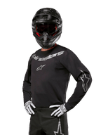 Zestaw Alpinestars 2024 Fluid Graphite, zawodnik motocrossowy w czarno-szarej koszulce z wyraźnymi szarymi logotypami, pasujące spodnie, czarny kask i biało-czarne rękawice, sportowy krój do wyścigów motocrossowych off-road