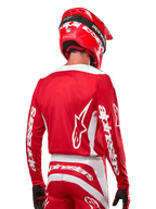 Kierowca motocrossowy w koszulce Alpinestars 2024 Fluid Lurv, kolor czerwono-biały, widok z tyłu ukazujący białe logo Alpinestars na plecach i branding na rękawach, w zestawie z dopasowanym czerwonym kaskiem i czarnymi rękawicami, profesjonalna odzież do wyścigów off-road