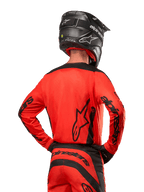 Motocyklista motocrossowy widoczny od tyłu w koszulce Alpinestars 2024 Fluid Lurv w kolorze jaskrawopomarańczowym i czarnym, z wyraźnym brandingiem Alpinestars na plecach i rękawach, w zestawie z dopasowanymi spodniami motocrossowymi i czarnym kaskiem, zaprojektowana do jazdy off-road.