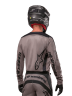 Motocyklista motocrossowy widziany od tyłu w koszulce Alpinestars 2024 Fluid Lurv w kolorze Mud Black, czarne logo Alpinestars na rękawach i plecach, czarny kask i dopasowany sprzęt do wyścigów terenowych