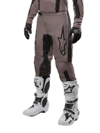 Alpinestars 2024 Fluid Lurv Pants, spodnie motocrossowe, kolor Mud Black i czarny, z logo Alpinestars, noszone z czarnymi rękawicami i białymi butami motocrossowymi, sportowy design do wyścigów terenowych