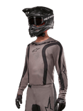 Alpinestars 2024 Fluid Lurv Jersey, koszulka motocrossowa w kolorze szarym i czarnym (Mud Black), model z długim rękawem, czarnymi zakrzywionymi pasami i logo, zestawiona z pasującymi spodniami i czarnym kaskiem motocrossowym, profesjonalny strój off-road