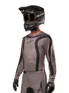 Alpinestars 2024 Fluid Lurv Jersey, koszulka motocrossowa w kolorze szarym i czarnym (Mud Black), model z długim rękawem, czarnymi zakrzywionymi pasami i logo, zestawiona z pasującymi spodniami i czarnym kaskiem motocrossowym, profesjonalny strój off-road