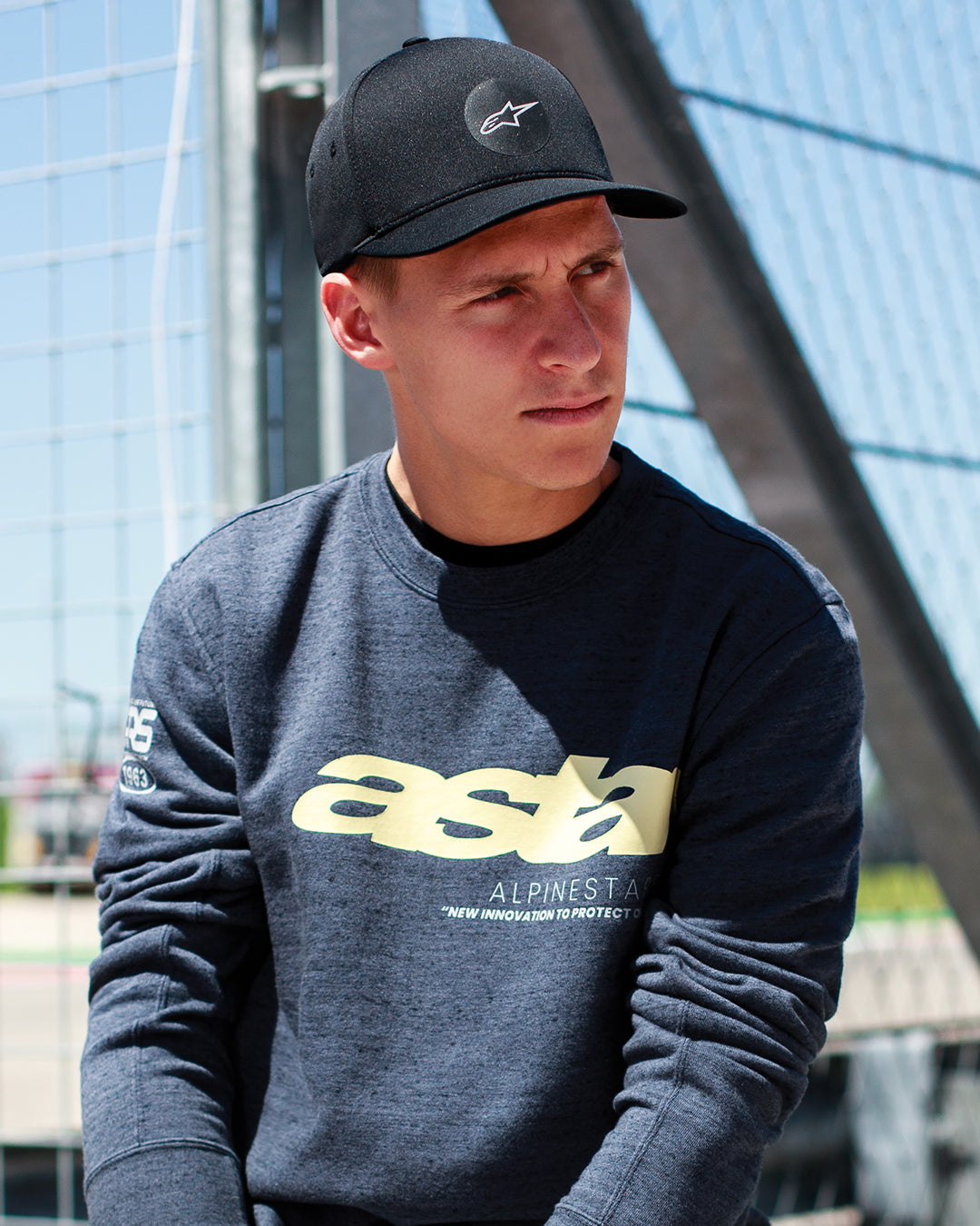 Fabio Quartararo ubrany w czarną czapkę Alpinestars Apex Hat, model o strukturalnej koronie z zakrzywionym daszkiem i okrągłym logo z gwiazdą, zestawiony z ciemnoszarą melanżową bluzą z żółtym brandingiem, codzienny styl inspirowany motorsportem.