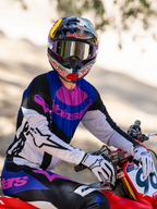 Motocross rider on a red dirt bike, Limited Edition HUNTA96 XXV Supertech Vision Goggles in purple and white with silver mirror lenses, matching purple black and white Alpinestars jersey and pants, Red Bull helmet, white gloves, coordinated professional motocross racing gear.