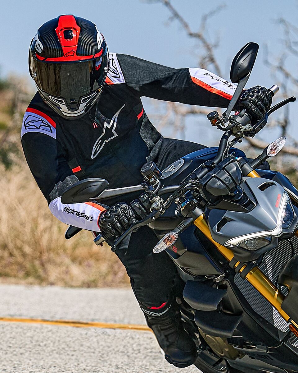 Motocyklista pochylający się w zakręcie na motocyklu typu naked, ubrany w kurtkę Alpinestars T-SPS Air v2 w kolorze czarnym z białymi i czerwonymi akcentami fluo, w czarno-czerwonym kasku i czarnych rękawicach; sportowa odzież tekstylna do jazdy miejskiej.