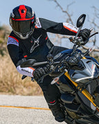 Motocyklista pochylający się w zakręcie na motocyklu typu naked, ubrany w kurtkę Alpinestars T-SPS Air v2 w kolorze czarnym z białymi i czerwonymi akcentami fluo, w czarno-czerwonym kasku i czarnych rękawicach; sportowa odzież tekstylna do jazdy miejskiej.