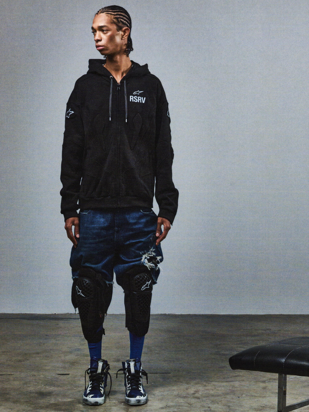 Alpinestars RSRV GP Hoodie, czarna bluza z kapturem, logo RSRV i gwiazdy, miejski styl motocyklowy, noszona z niebieskimi szortami i ochraniaczami na kolana