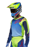 Koszulka Alpinestars Racer Hoen 2024, koszulka motocrossowa, kolory żółty fluo i granatowy z czerwonymi logotypami, noszona z pasującym żółto-niebiesko-czerwonym kaskiem i niebieskimi rękawicami, wyrazisty strój do jazdy terenowej