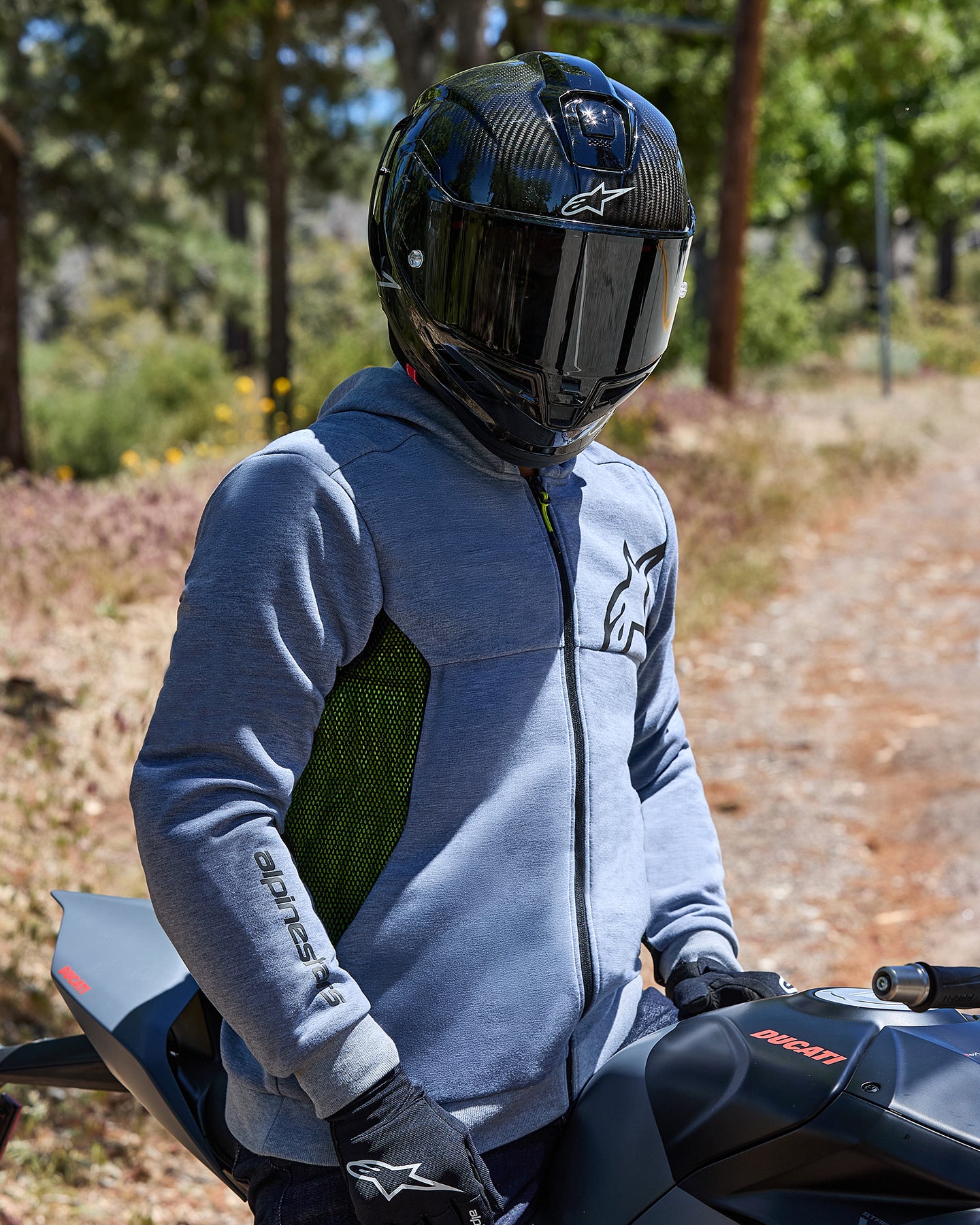 Motocyklista ubrany w bluzę Alpinestars Chrome Air Sport Hoodie w kolorze Gray Heather, z boczno-siatkowymi panelami w kolorze limonkowym i czarnym logo, w czarnym kasku z włókna węglowego i czarnych rękawicach, siedzący na motocyklu Ducati w plenerze na drodze