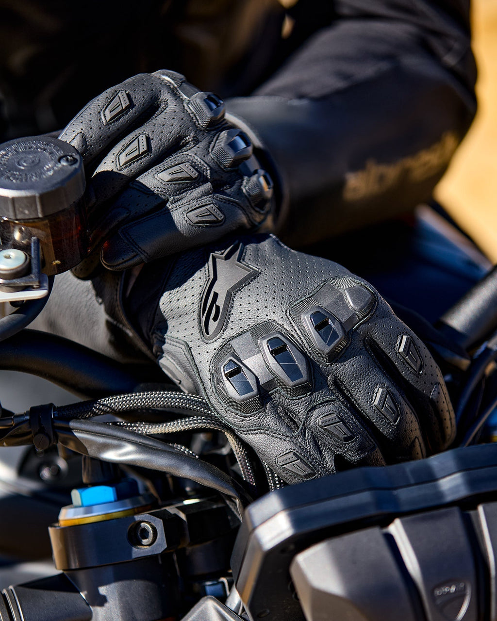 Alpinestars SP X 7 Gloves, czarne perforowane skórzane rękawice motocyklowe z ochraniaczami kostek TPU i ślizgaczami palców, widoczne na dłoniach motocyklisty trzymającego kierownicę motocykla