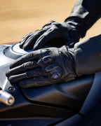 Alpinestars SP X 7 Gloves, czarne, krótkie rękawice motocyklowe z perforowanej skóry, dłonie oparte na ciemnym baku motocykla, widoczne ochraniacze kostek TPU o podwójnej gęstości i tłoczone logo, przeznaczone do jazdy sportowej i szosowej.