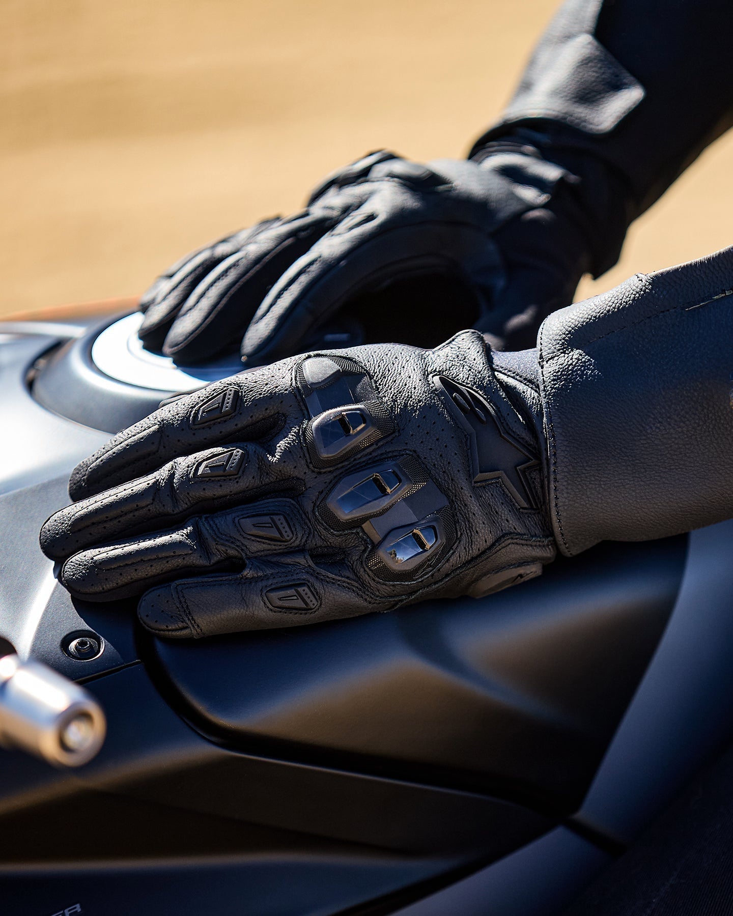 Alpinestars SP X 7 Gloves, czarne, krótkie rękawice motocyklowe z perforowanej skóry, dłonie oparte na ciemnym baku motocykla, widoczne ochraniacze kostek TPU o podwójnej gęstości i tłoczone logo, przeznaczone do jazdy sportowej i szosowej.
