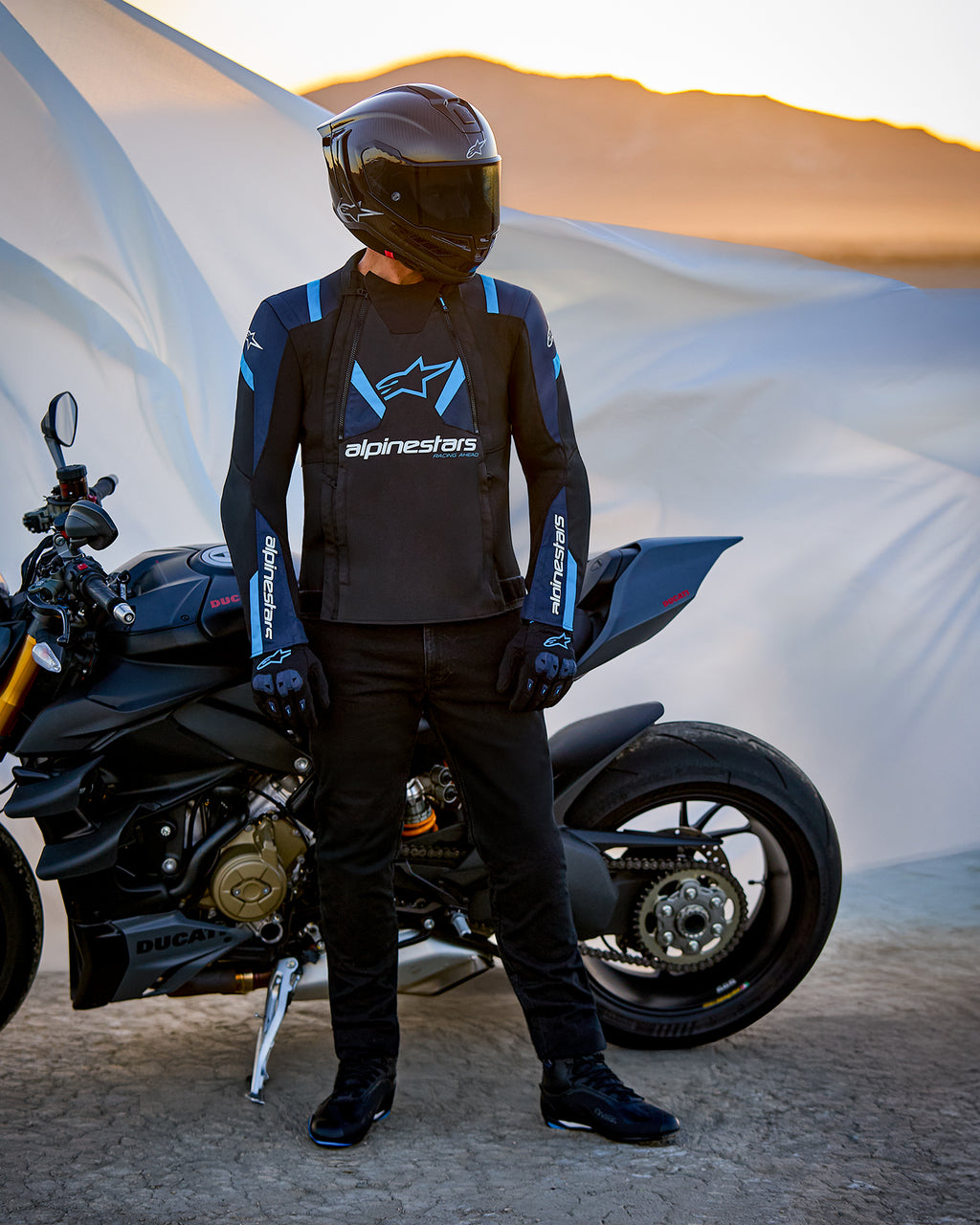 Alpinestars T-Stunt Air Jacket, sportowa kurtka tekstylna, czarna, granatowa i jasnoniebieska, motocyklista stojący obok motocykla Ducati, kask z włókna węglowego i czarny strój motocyklowy, pustynny krajobraz o zachodzie słońca