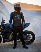 Alpinestars T-Stunt Air Jacket, sportowa kurtka tekstylna, czarna, granatowa i jasnoniebieska, motocyklista stojący obok motocykla Ducati, kask z włókna węglowego i czarny strój motocyklowy, pustynny krajobraz o zachodzie słońca