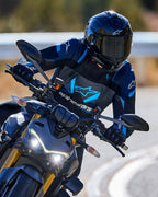 Motocyklista w pochyleniu na zakręcie, ubrany w kurtkę Alpinestars T-Stunt Air w kolorach czarnym, ciemnoniebieskim i jasnoniebieskim, czarny kask i rękawice, dynamiczne ujęcie na drodze asfaltowej, sportowa odzież motocyklowa