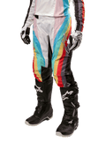 Alpinestars Stella Techstar 2024, damskie spodnie motocrossowe, czarne wielokolorowe z ukośnymi czerwonymi, żółtymi i niebieskimi pasami, widoczne z czarnymi butami i białymi rękawicami, z czarnymi panelami ochronnymi na kolanach i wyrazistym logo do wyścigów off-road
