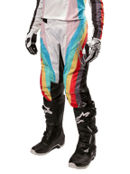 Alpinestars Stella Techstar 2024, damskie spodnie motocrossowe, czarne wielokolorowe z ukośnymi czerwonymi, żółtymi i niebieskimi pasami, widoczne z czarnymi butami i białymi rękawicami, z czarnymi panelami ochronnymi na kolanach i wyrazistym logo do wyścigów off-road