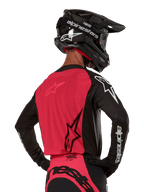 Alpinestars 2024 Supertech Ward Jersey, koszulka motocrossowa, czarno-czerwona (Red Berry), widok z tyłu prezentujący czerwony panel z siatki i czarne rękawy z białym brandingiem, zestawiona z czarnym kaskiem z włókna węglowego i rękawicami, sportowy krój wyścigowy do jazdy terenowej