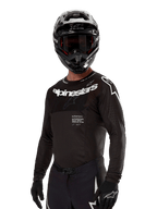 Koszulka Alpinestars Supertech Ward 2024, koszulka motocrossowa, czarno-biała, techniczna odzież z długim rękawem i wyraźnym logo Alpinestars, widoczna z dopasowanym kaskiem i rękawicami, sportowy krój do jazdy off-road