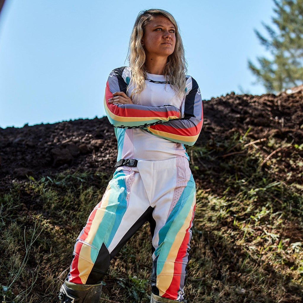 Motocyklistka motocrossowa w spodniach Alpinestars Stella Techstar 2024 i pasującej koszulce, wzór Black Multicolor z białą bazą oraz niebieskimi, żółtymi i czerwonymi pasami, stojąca z założonymi rękami na torze ziemnym, profesjonalny strój wyścigowy off-road