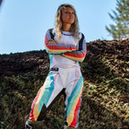 Motocyklistka motocrossowa w spodniach Alpinestars Stella Techstar 2024 i pasującej koszulce, wzór Black Multicolor z białą bazą oraz niebieskimi, żółtymi i czerwonymi pasami, stojąca z założonymi rękami na torze ziemnym, profesjonalny strój wyścigowy off-road