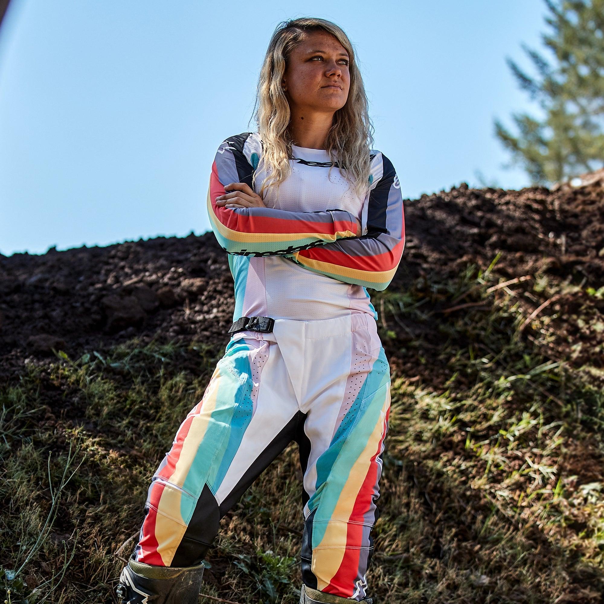 Motocyklistka motocrossowa w spodniach Alpinestars Stella Techstar 2024 i pasującej koszulce, wzór Black Multicolor z białą bazą oraz niebieskimi, żółtymi i czerwonymi pasami, stojąca z założonymi rękami na torze ziemnym, profesjonalny strój wyścigowy off-road