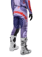 Alpinestars Techstar Ocuri Pants 2024, spodnie motocrossowe, fioletowe z intensywnymi pomarańczowymi i ciemnoniebieskimi akcentami, duże pomarańczowe logo Alpinestars z tyłu, białe buty motocyklowe, sportowy krój wyścigowy