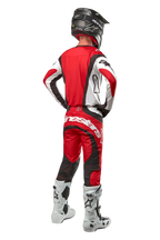 Alpinestars 2024 Techstar Ocuri Pants, spodnie motocrossowe, kolor Mars Red White Black, widok z tyłu na zawodnika motocrossowego w pełnym stroju wyścigowym, w tym pasującą bluzę, czarny kask oraz biało-czarne buty, podkreślający branding Alpinestars i trwałą konstrukcję materiału.