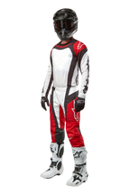 Zawodnik motocrossowy w pełnym stroju Alpinestars, spodnie Techstar Ocuri 2024 w kolorze Mars Red White Black, pasująca koszulka, czarny kask z goglami, czarno-białe rękawice i białe buty motocrossowe, profesjonalny zestaw off-road