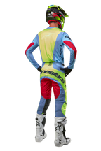Kierowca motocrossowy widoczny od tyłu w spodniach i pasującej koszulce Techstar Ocuri 2024, kolory jasnoniebieski, neonowy żółty i czerwony Red Berry, biało-czarne buty, czarny kask z neonowymi akcentami, dopasowany strój Alpinestars do wyścigów off-road