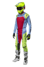 Motocyklista motocrossowy w spodniach Alpinestars Techstar Ocuri 2024 w kolorach jasnoniebieskim, neonowym żółtym i jagodowym czerwonym, dopasowany zestaw wyścigowy z pasującą bluzą, neonowożółtym kaskiem i białymi butami, widok z przodu profesjonalnego stroju terenowego