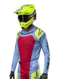 Alpinestars 2024 Techstar Ocuri Jersey, koszulka motocrossowa, jasnoniebieska z czerwonymi i neonowożółtymi akcentami, noszona z pasującym neonowożółtym kaskiem i czarnymi goglami, czarne logo Alpinestars na klatce piersiowej i ramionach, sportowy design inspirowany wyścigami do jazdy w terenie