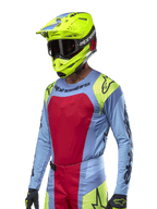 Alpinestars 2024 Techstar Ocuri Jersey, koszulka motocrossowa, jasnoniebieska z czerwonymi i neonowożółtymi akcentami, noszona z pasującym neonowożółtym kaskiem i czarnymi goglami, czarne logo Alpinestars na klatce piersiowej i ramionach, sportowy design inspirowany wyścigami do jazdy w terenie