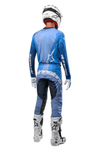 Motocyklista motocrossowy widziany od tyłu, ubrany w spodnie Alpinestars 2024 Techstar Pneuma w kolorze ciemnogranatowym i jasnoniebieskim, zestawione z pasującą niebieską koszulką, białym kaskiem, białymi rękawicami i białymi butami, z wyraźnym brandingiem Alpinestars na siedzeniu i rękawach.