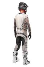 Alpinestars Techstar Pneuma Pants 2024, spodnie motocrossowe, kolorystyka Dark Sand i Iron Dust Gray, widok z tyłu na motocykliście w pasującej koszulce, biało-czarnych butach i czarnym kasku, z wyraźnym brandingiem Alpinestars na siedzeniu i wzmocnionymi ciemnymi panelami