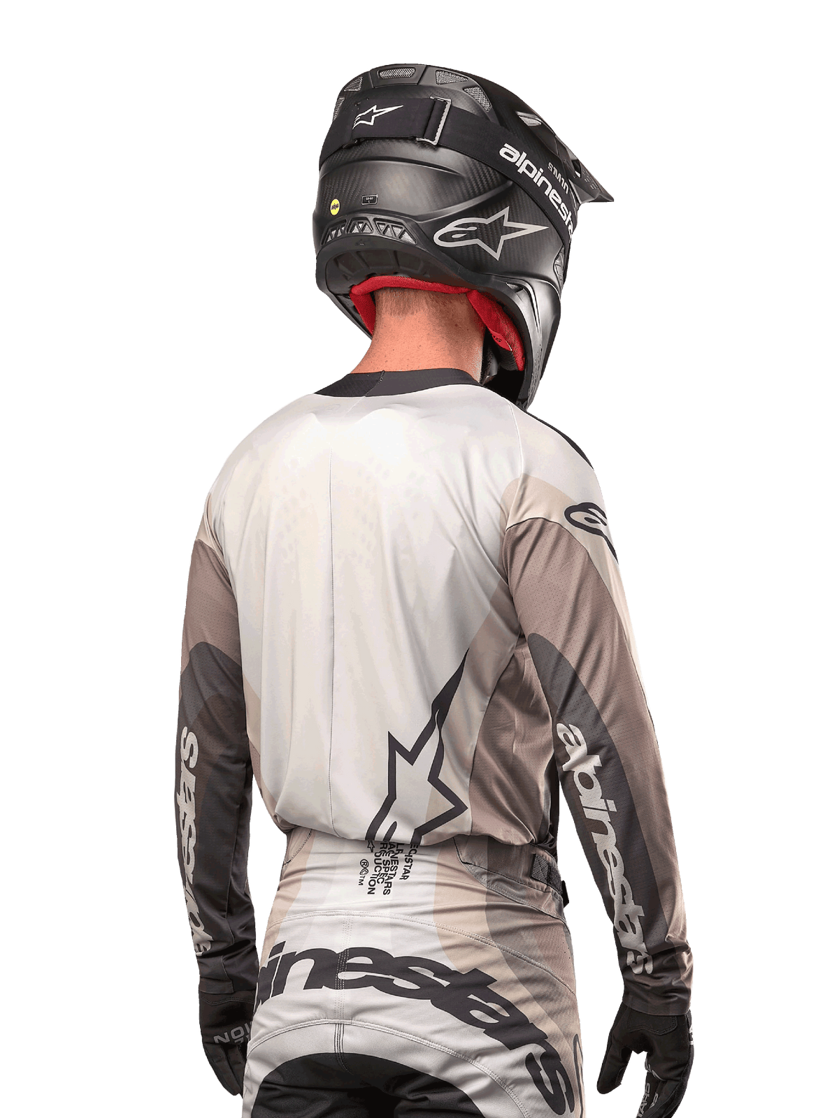 Kierowca motocrossowy widoczny od tyłu, koszulka Alpinestars 2024 Techstar Pneuma w kolorze Dark Sand Iron Dust Gray, czarny kask o karbonowym wykończeniu, duże logo gwiazdy na plecach i branding Alpinestars na rękawach.