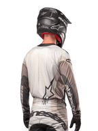 Kierowca motocrossowy widoczny od tyłu, koszulka Alpinestars 2024 Techstar Pneuma w kolorze Dark Sand Iron Dust Gray, czarny kask o karbonowym wykończeniu, duże logo gwiazdy na plecach i branding Alpinestars na rękawach.