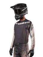 Alpinestars Techstar Pneuma Jersey 2024, koszulka motocrossowa, ciemnopiaskowa i szara (Iron Dust Gray), techniczna koszulka z długim rękawem, laserowo wycinane perforacje i białe logo, noszona z pasującymi spodniami i czarnym kaskiem, sportowy krój off-road
