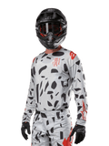 Alpinestars 2024 Techstar Rantera Jersey, koszulka motocrossowa, kolor Haze Gray Camo z czarnymi abstrakcyjnymi wzorami i czerwonymi akcentami, model z długim rękawem noszony z pasującym czarnym kaskiem i rękawicami, techniczna odzież do wyścigów terenowych