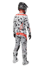 Motocyklista motocrossowy widoczny od tyłu, ubrany w spodnie Alpinestars 2024 Techstar Rantera, kolor Haze Gray Camo z czarnymi plamami i czerwonymi logotypami, pasująca koszulka, czarny kask i białe buty, kompletny strój wyścigowy off-road