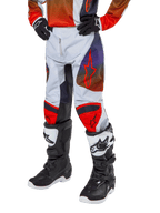 Alpinestars 2024 Youth Racer Hoen Pants, spodnie motocrossowe, jasnoszare z jaskrawopomarańczowymi i czarnymi akcentami, z czerwonym logo gwiazdy na udach i czerwonymi panelami na kolanach, noszone z czarno-białymi butami, wytrzymała konstrukcja do wyścigów terenowych