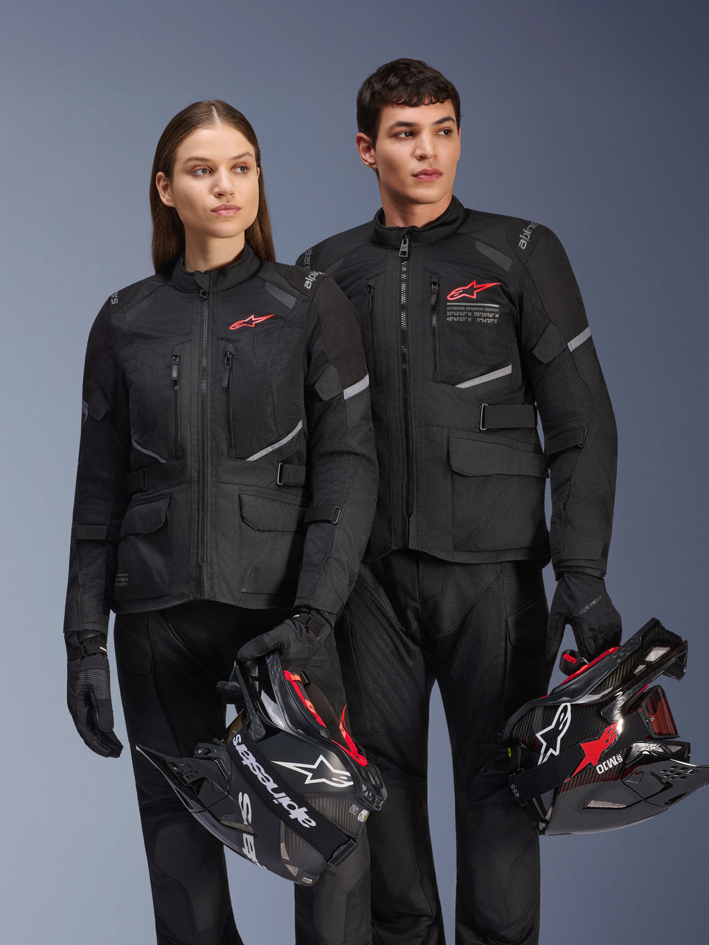 Mężczyzna i kobieta w kurtkach Alpinestars Andes Air Drystar®, czarna odzież adventure touring z panelami z siatki i odblaskowymi detalami, pasujące czarne spodnie i rękawice, trzymający kaski motocrossowe, zdjęcie studyjne profesjonalnej odzieży motocyklowej